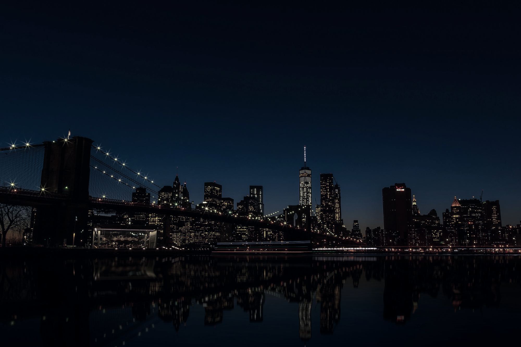 A suspension bridge lit at night, with a city skyline beyond and its lights on the water